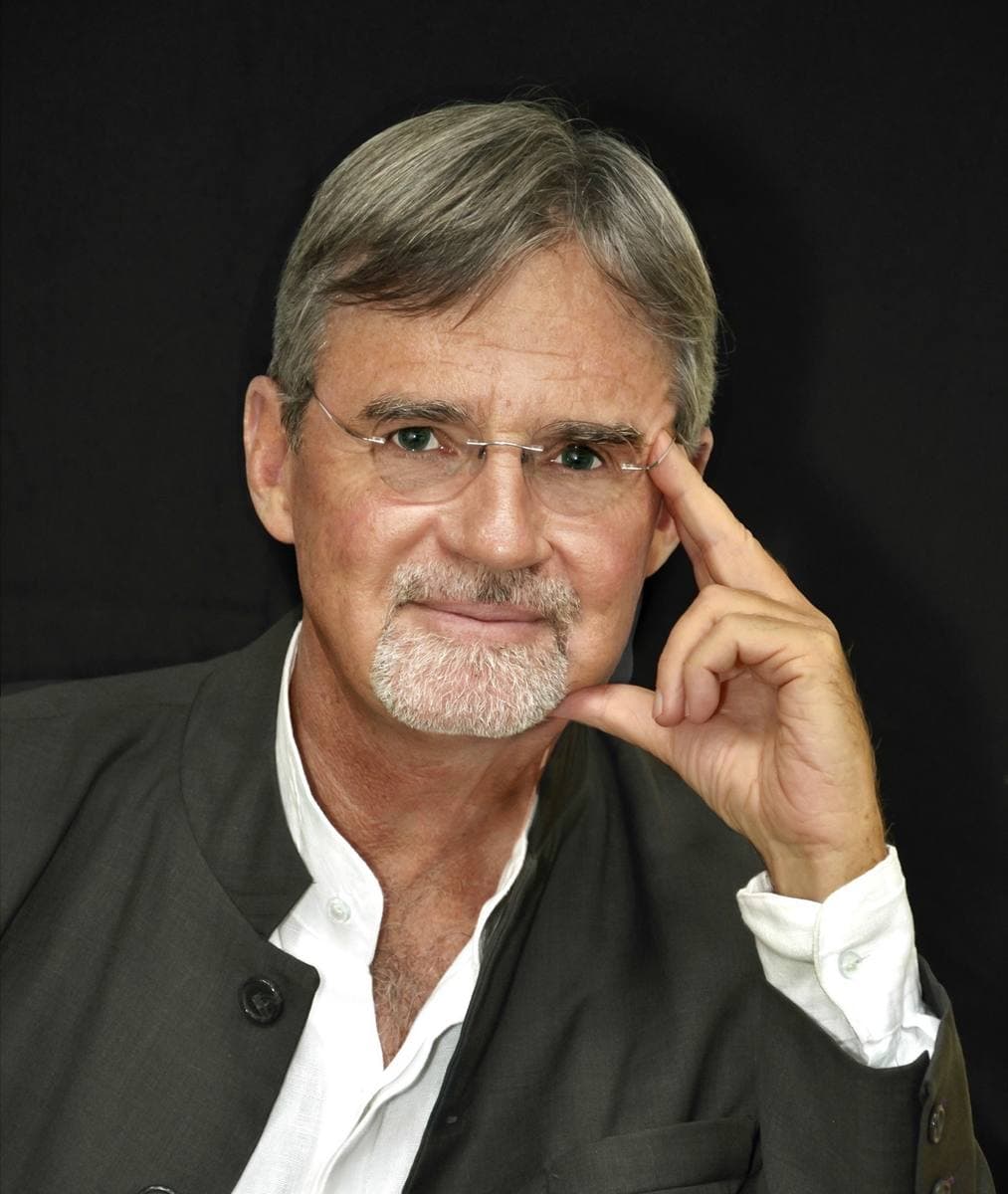 Kai Bird with grey hair and beard, resting his chin on his hand against a dark background