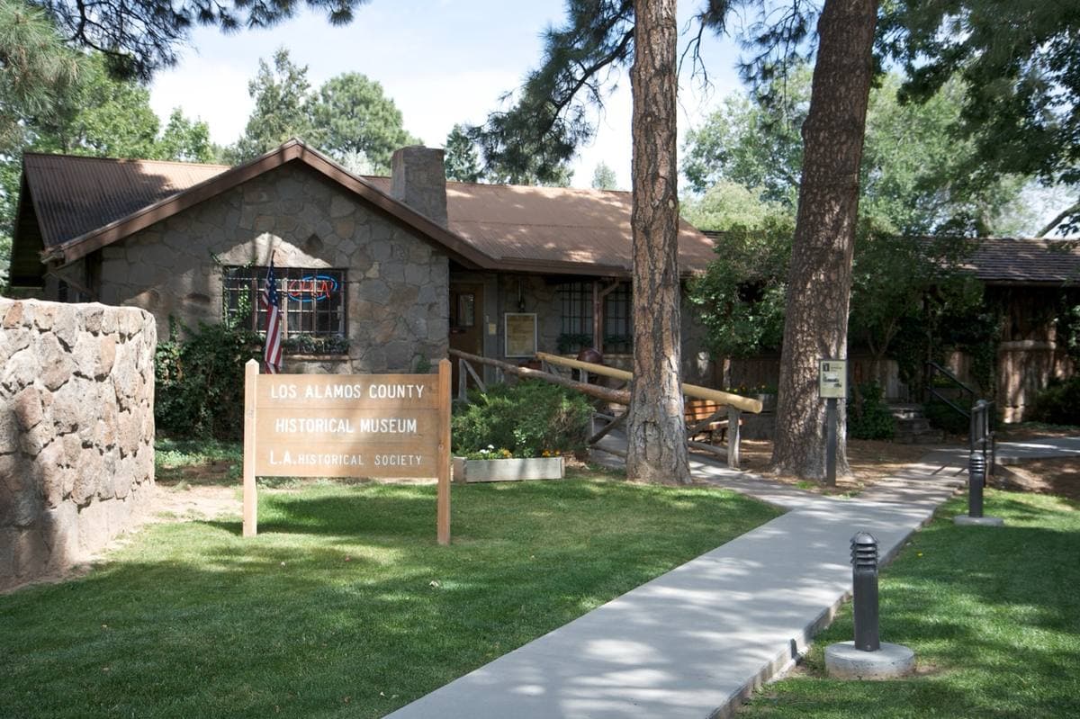 A low stone and log building behind a lawn, with a wooden sign by the path