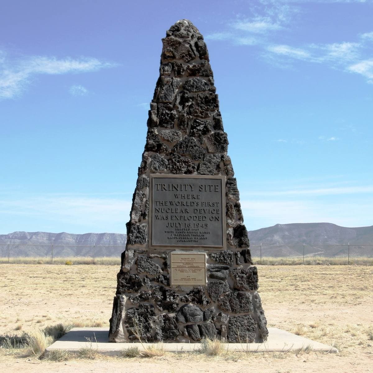 A dark stone obelisk with a black plaque, standing in flat desert under a blue sky