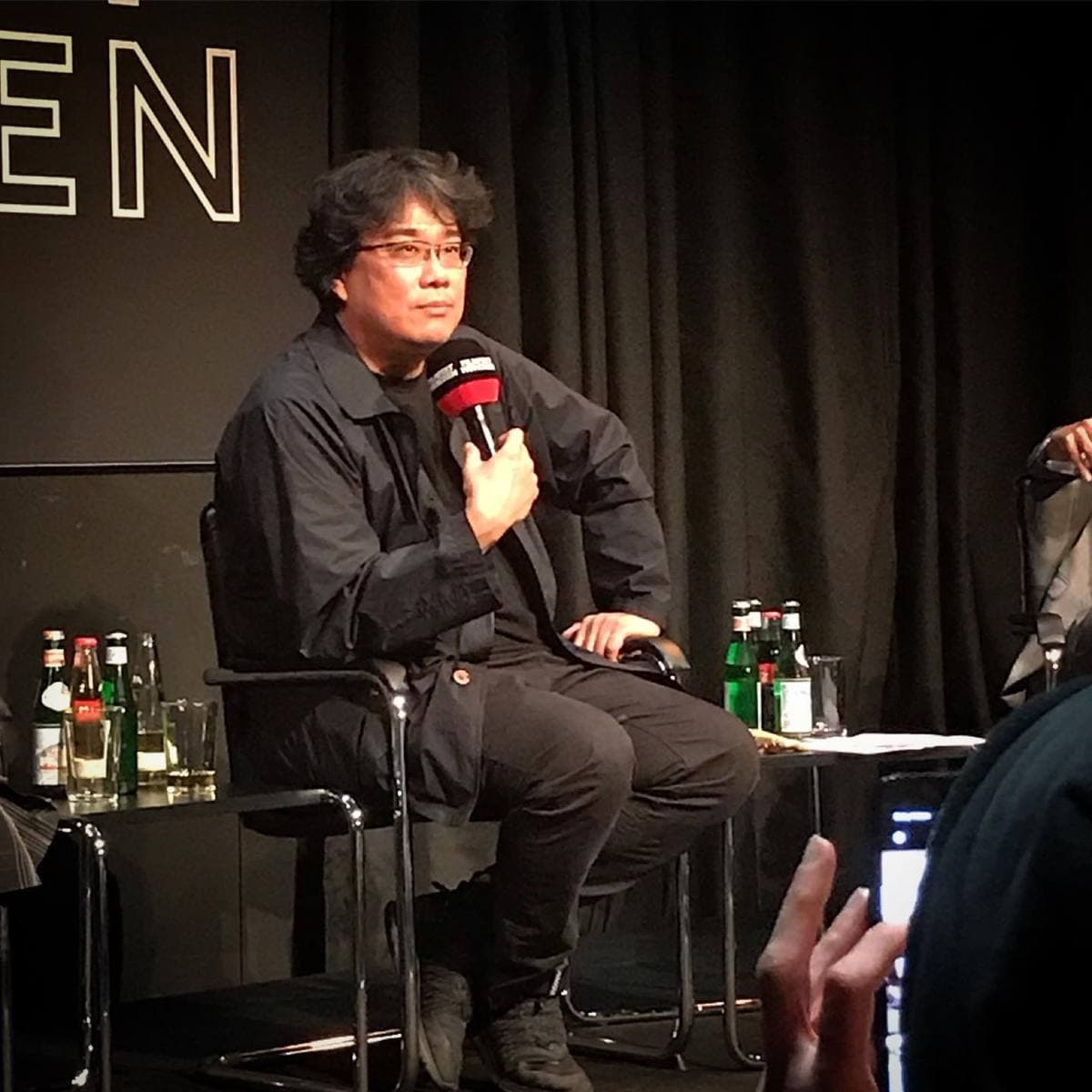 Colour photograph of Bong Joon-ho in glasses and a dark jacket, seated with a microphone