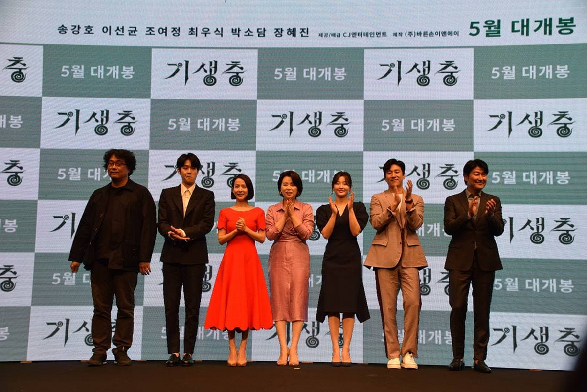Colour photograph of seven people standing in a line on a stage, applauding, in front of a green and white screen