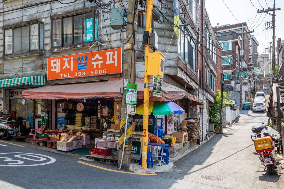 Colour photograph of a corner shop with an orange sign, a parasol and boxes of fruit outside, a lane rising behind it