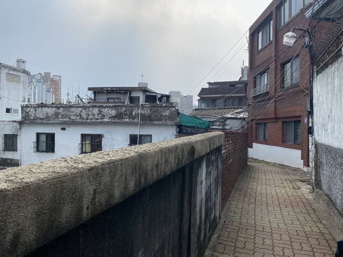 Colour photograph of a narrow paved lane along a concrete wall, low houses and rooftops below