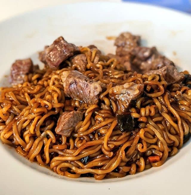 Colour photograph of a bowl of dark glossy noodles topped with pieces of beef