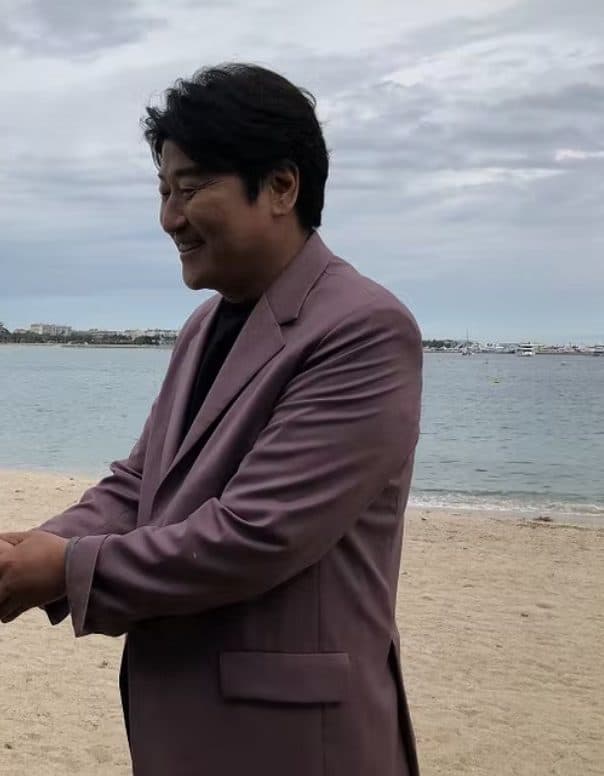 Colour photograph of Song Kang-ho in a mauve suit on a beach, smiling