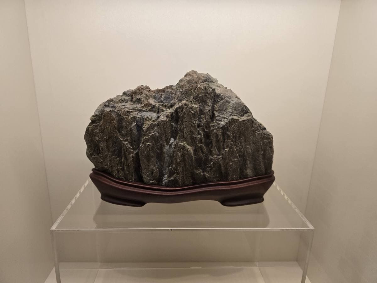 Colour photograph of a dark craggy stone like a small mountain on a carved wooden stand in a glass case