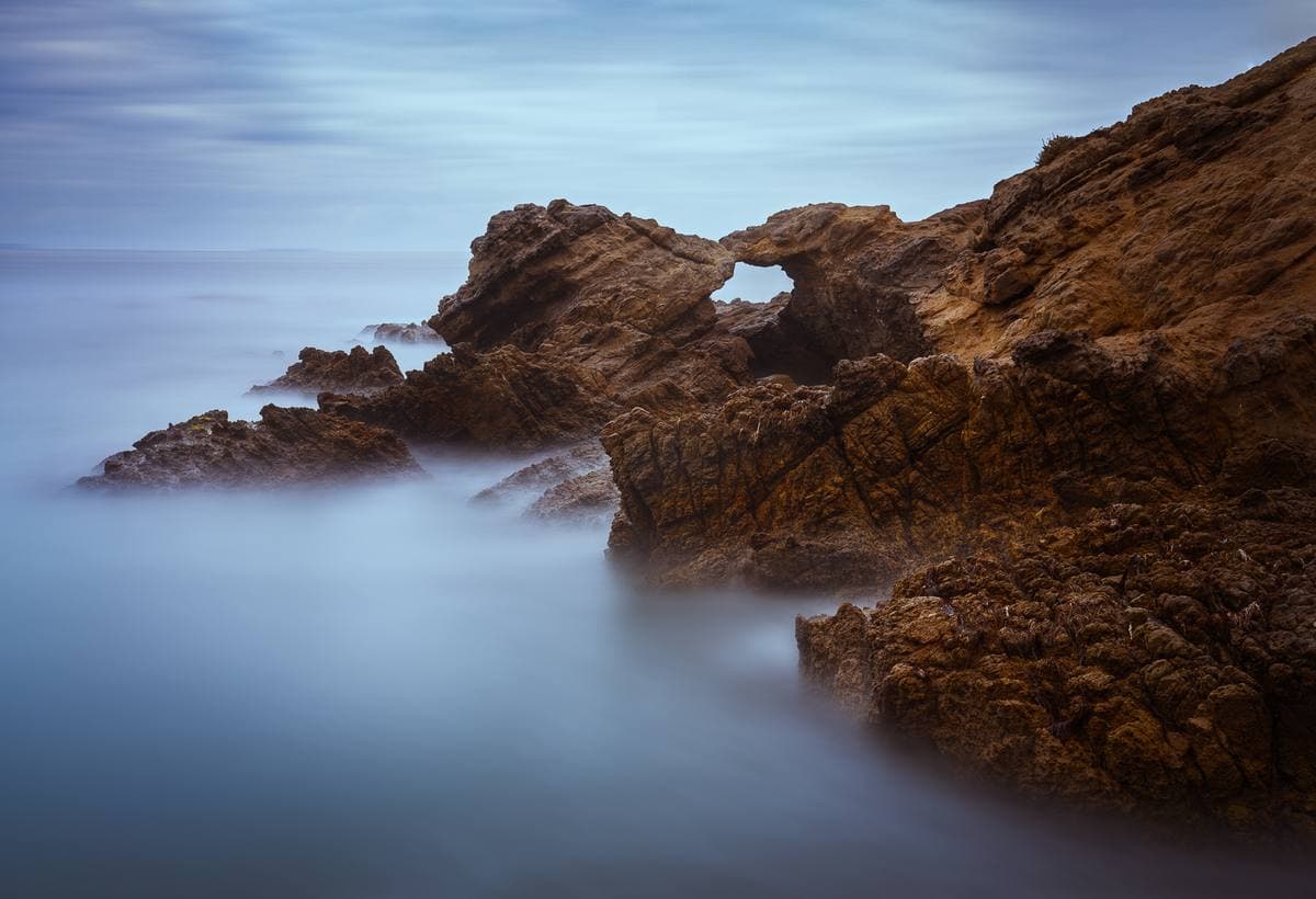 Brown sandstone rocks with an arch, the sea smoothed to mist by a long exposure