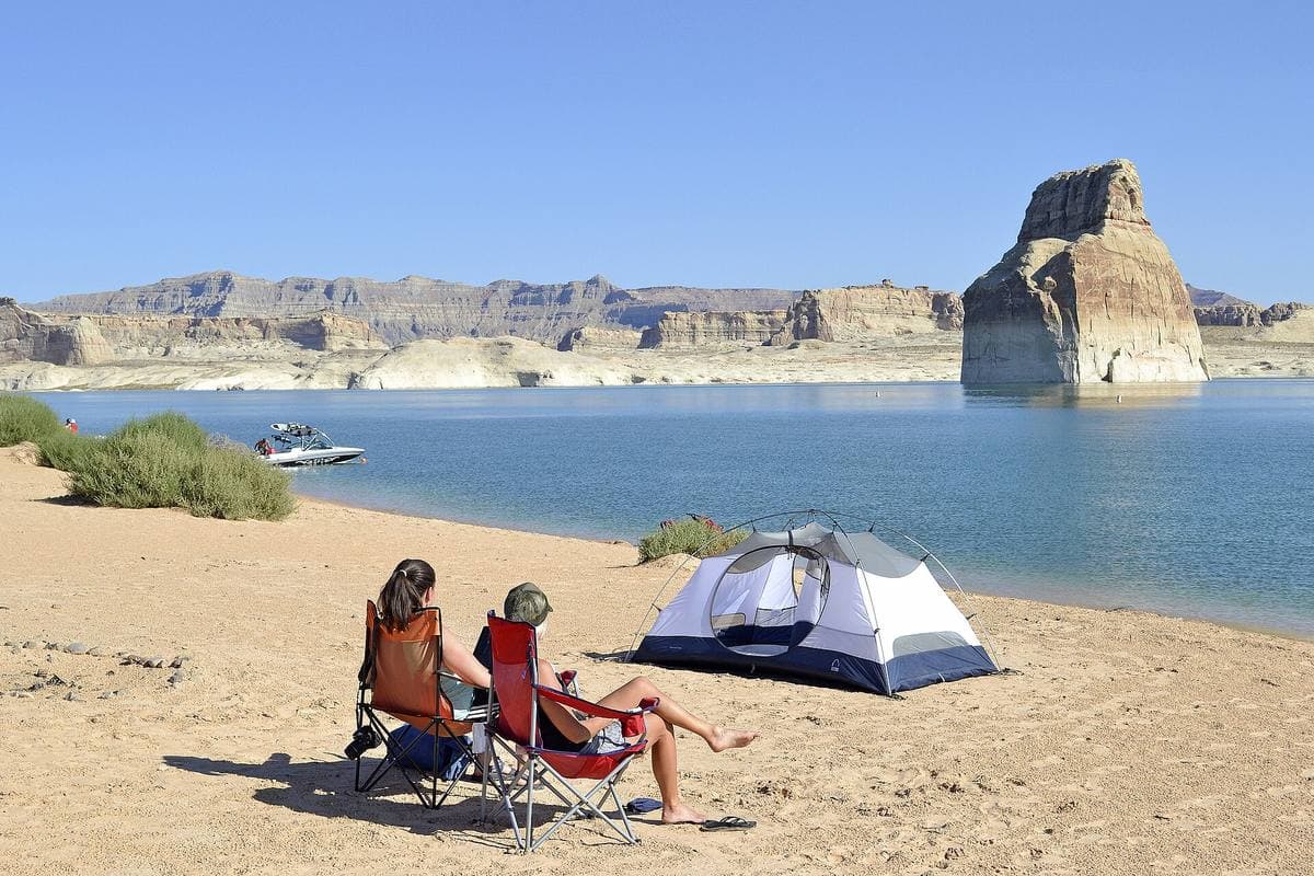Two people in camp chairs by a tent on a sandy shore, a lone rock tower in the lake