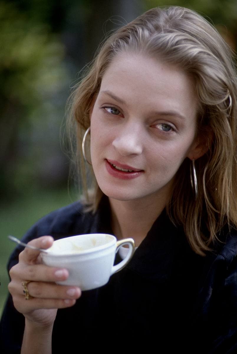 Uma Thurman, aged 24, resting her chin on her hands and smiling, with hoop earrings