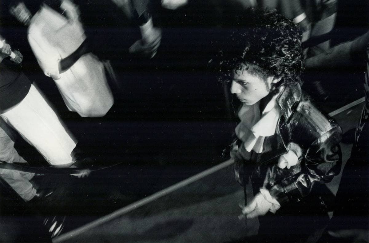 Black-and-white photograph of a man with curly dark hair in a ruffled shirt and studded jacket, moving through a blurred crowd