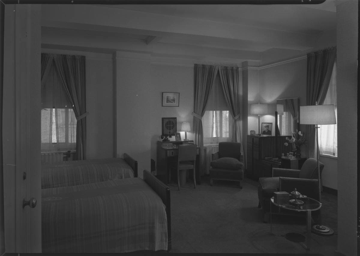 A 1940s hotel room with twin beds, armchairs and tall curtained windows