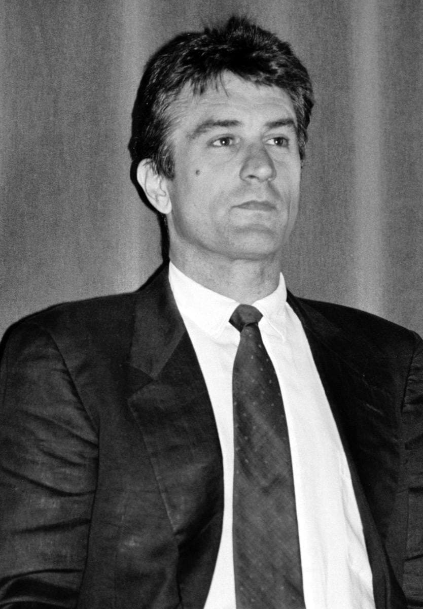 Robert De Niro, dark hair swept back, in a dark suit and white shirt, in front of a curtain