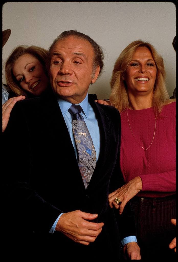 Jake LaMotta, grey-haired, in a dark suit and patterned tie, between two smiling blonde women