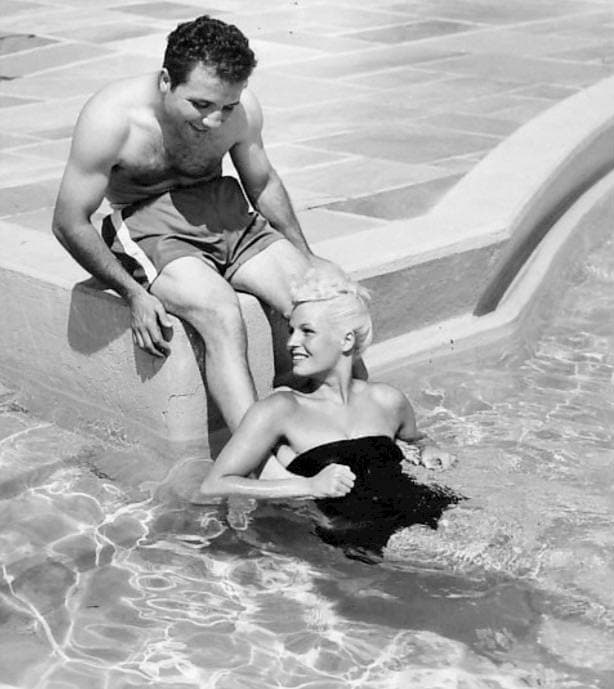 Jake LaMotta, bare-chested, sits on the edge of a pool while Vickie, in a black swimsuit, leans on the side in the water