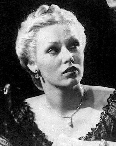 Cathy Moriarty, platinum hair pinned up, in a lace dress and a necklace, looking off to the side