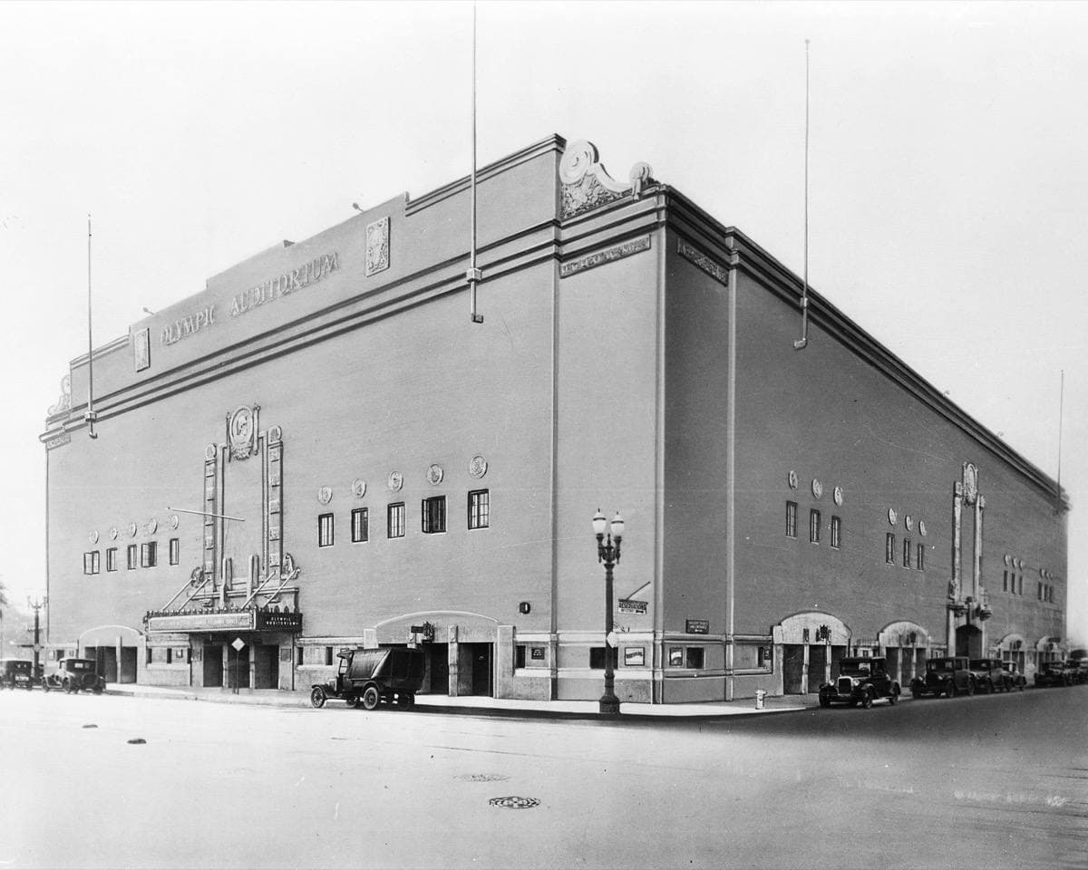 A long, plain arena building with a decorated entrance on a downtown street corner, old cars parked outside