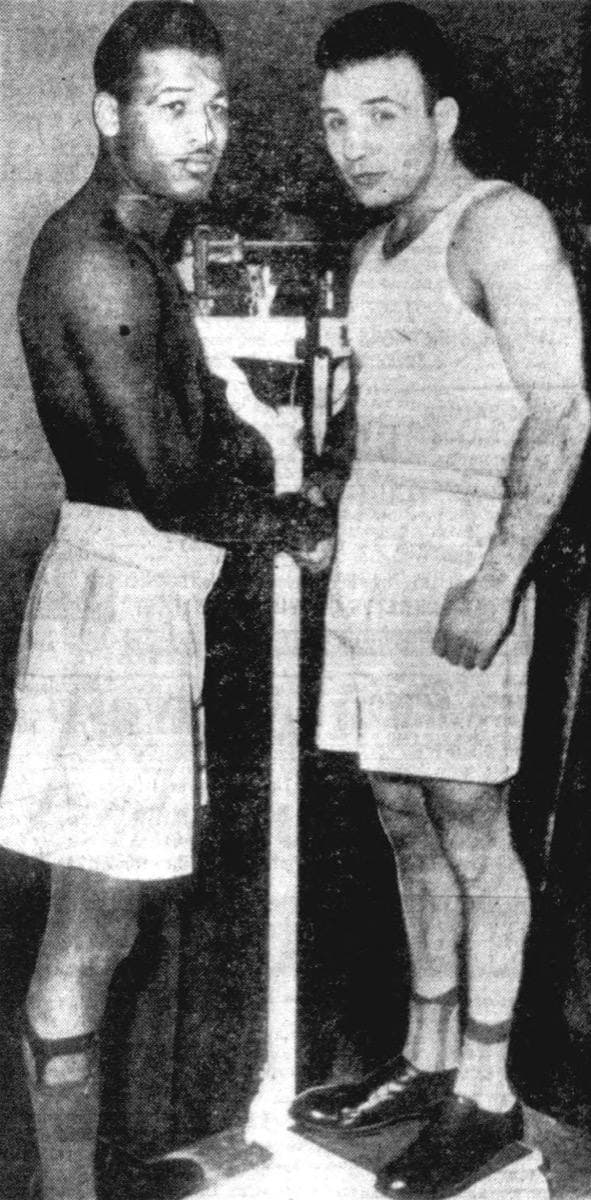 Two boxers in trunks stand either side of a tall balance-beam scale