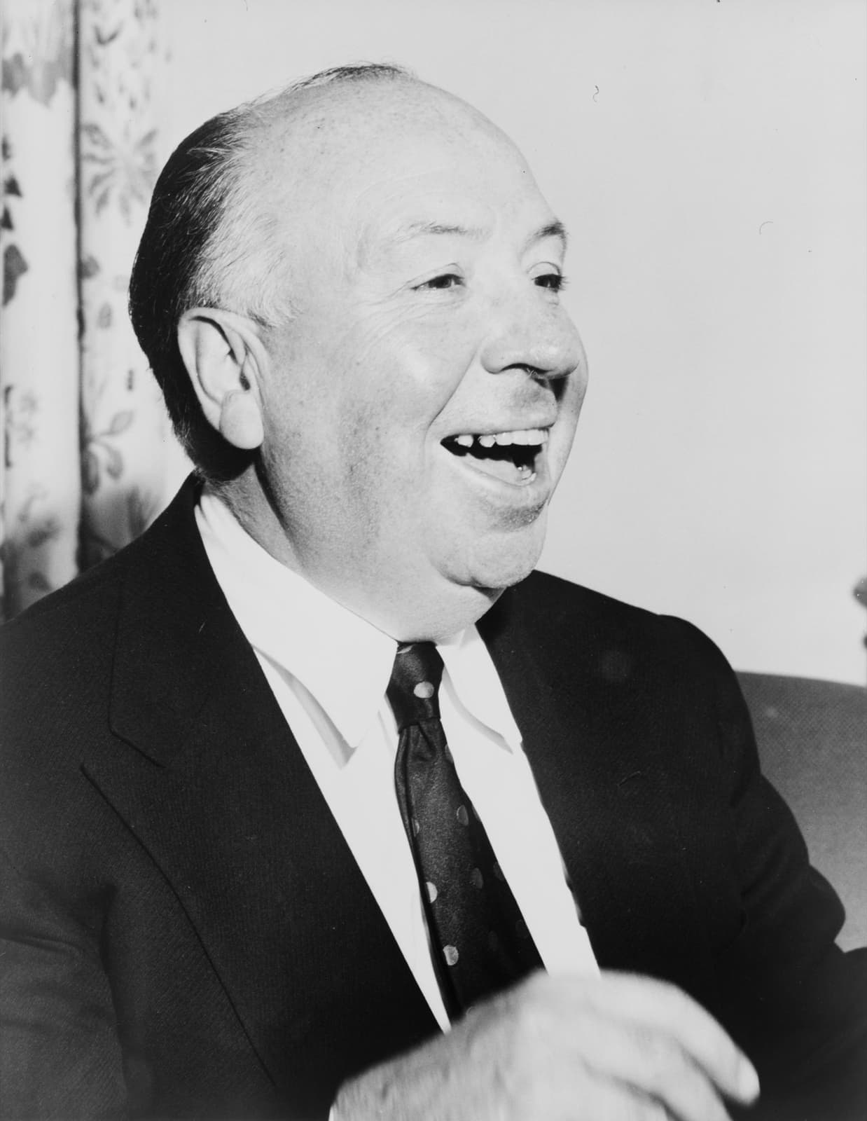 Alfred Hitchcock laughing, head and shoulders, in a dark suit and tie
