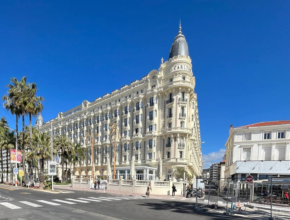 A long white belle-époque hotel with two domes behind palm trees
