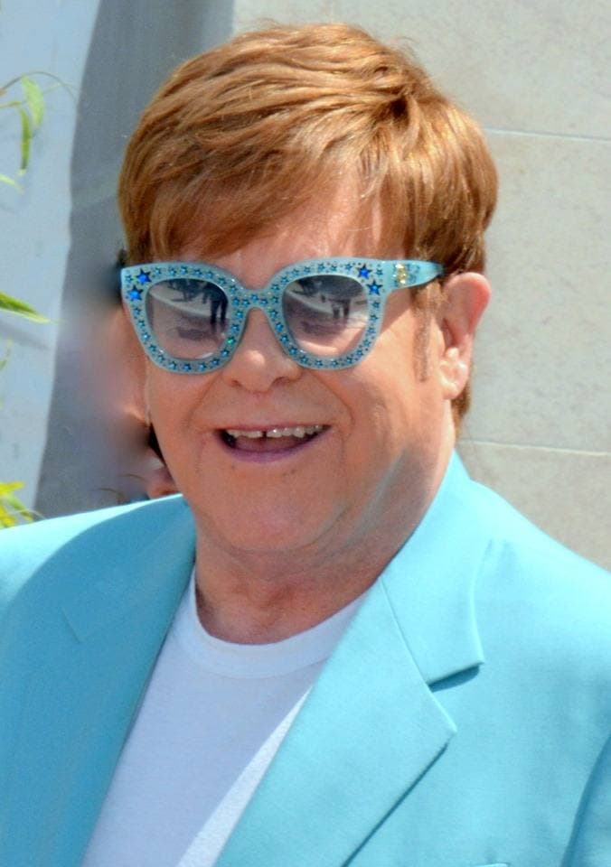 Elton John smiling in a pale blue jacket and jewelled blue glasses