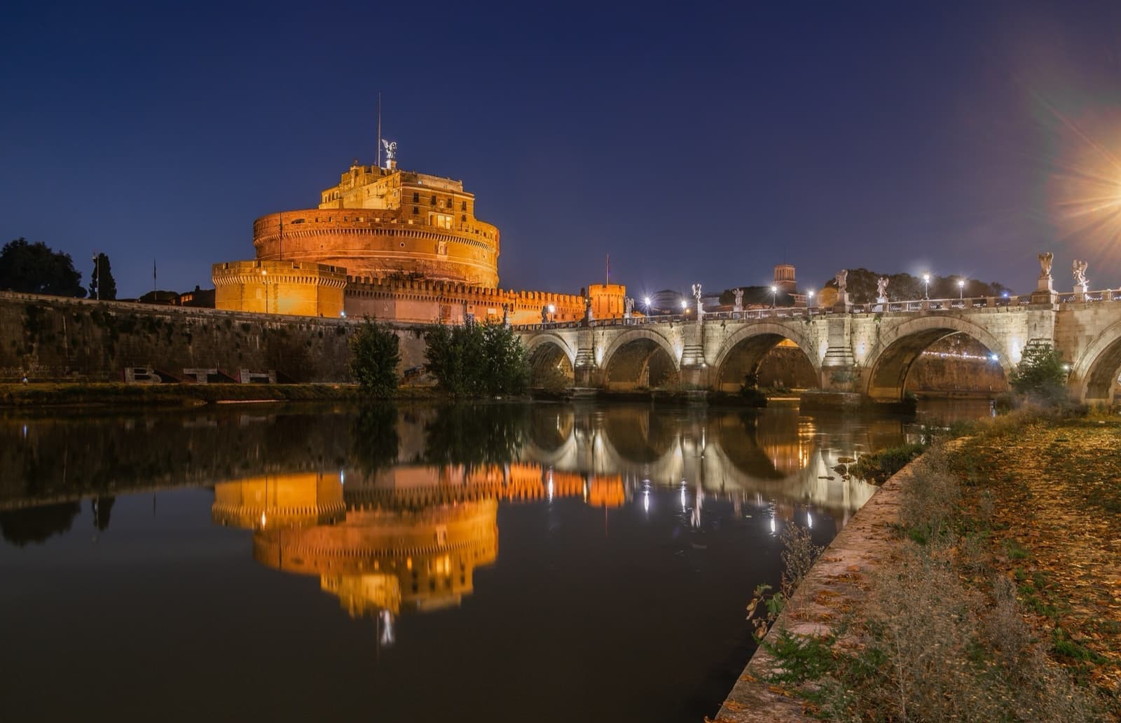 A floodlit bridge with statues leading to a round castle at night