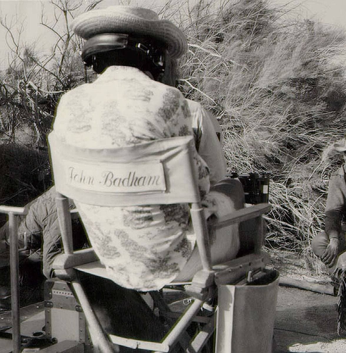 A man in a patterned shirt and a hat seen from behind in a canvas director’s chair with his name on the back, actors in the desert scrub beyond