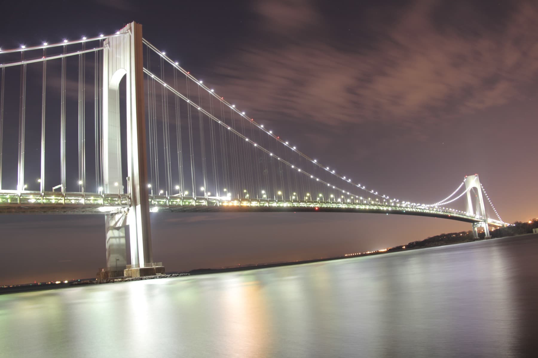 A long suspension bridge strung with lights across dark water at night, its tower lit on the left