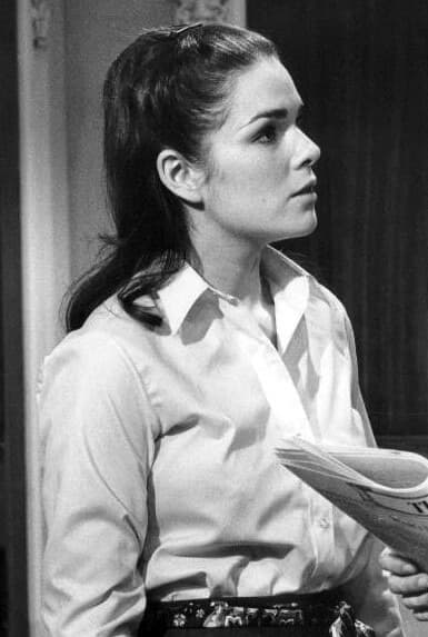 A young woman with long dark hair tied back, in a white collared shirt, looking to the side