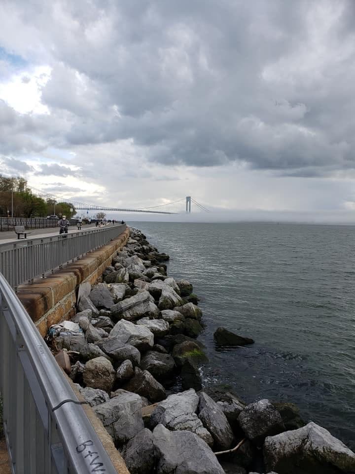 A railed waterside path above boulders, with a bridge faint in fog far along the shore