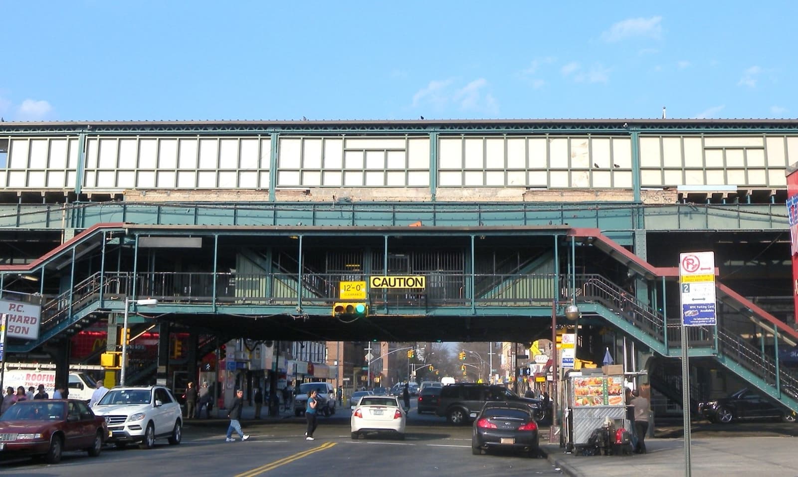 A green steel elevated railway station spanning a street, with stairs down to the pavement and cars below
