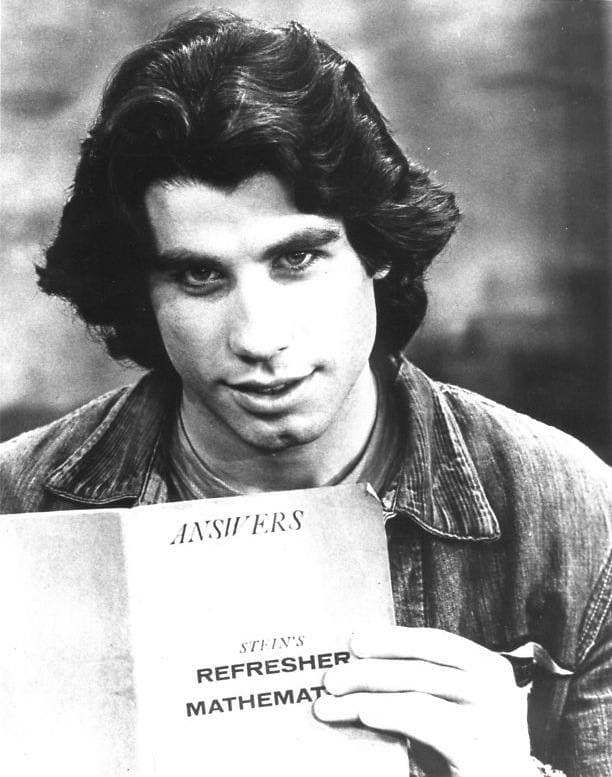 A young man with thick dark hair in a denim shirt, smiling and holding up a sheet of paper