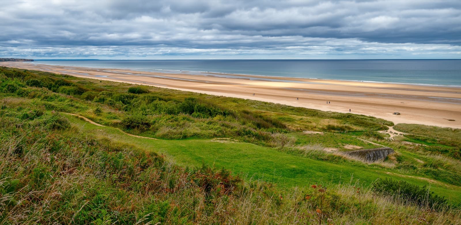 A long sandy beach and grassy bluff seen from high ground, the sea on the right