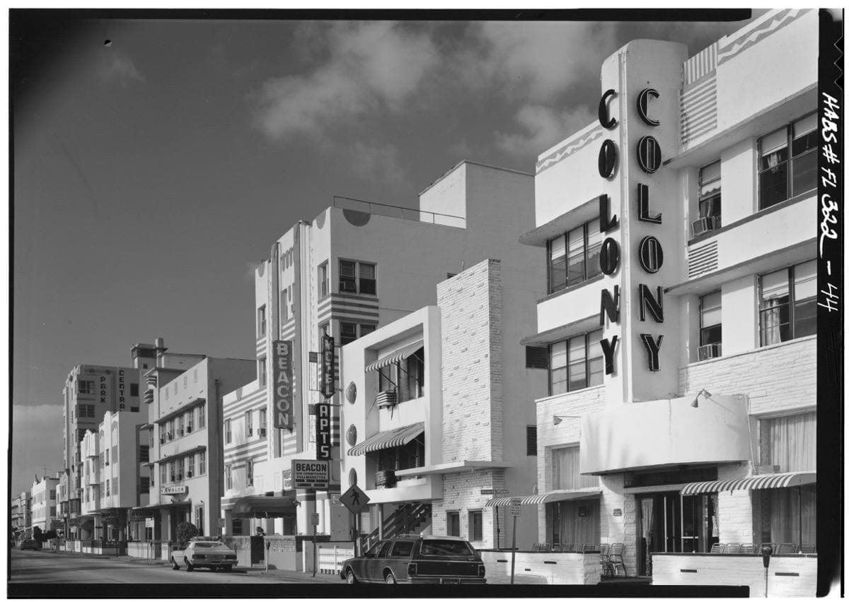 A black-and-white street of white Art Deco hotels with a tall vertical name fin reading Colony