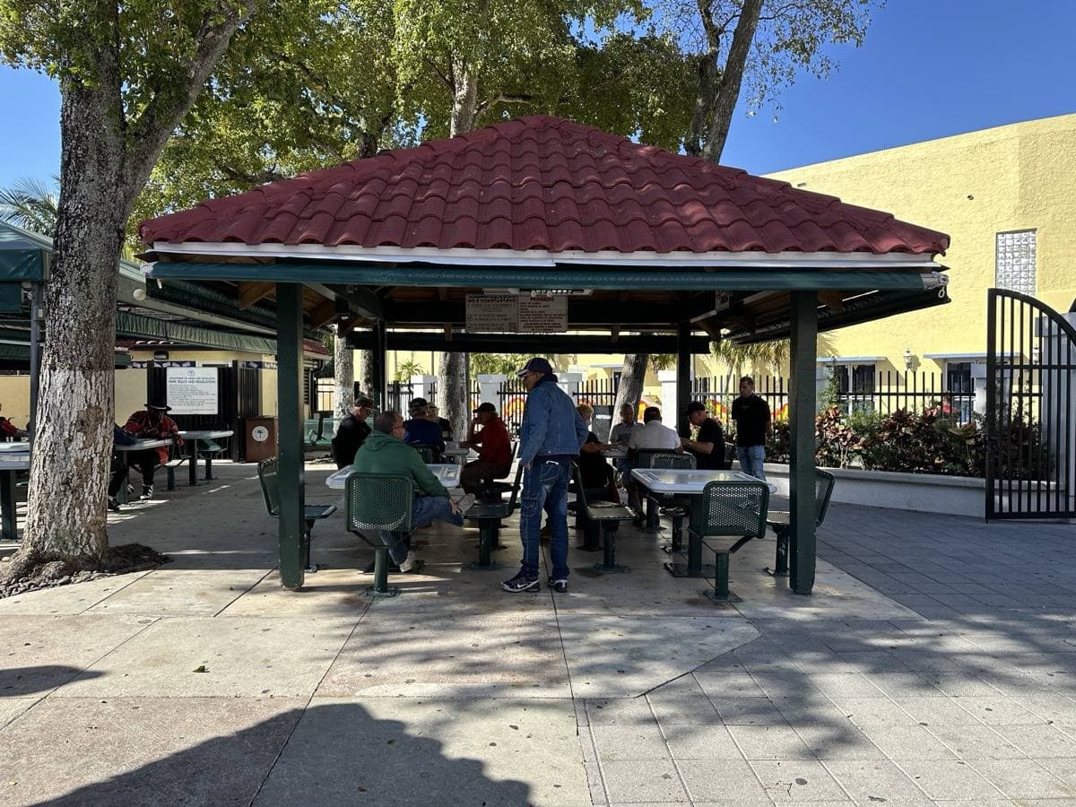 Men at tables under an open tiled pavilion, playing dominoes in the shade