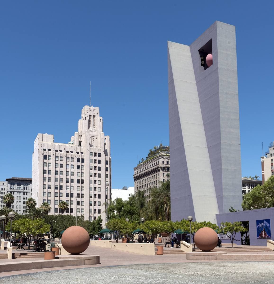 A plaza with large orange spheres, a tall purple tower and older downtown buildings behind