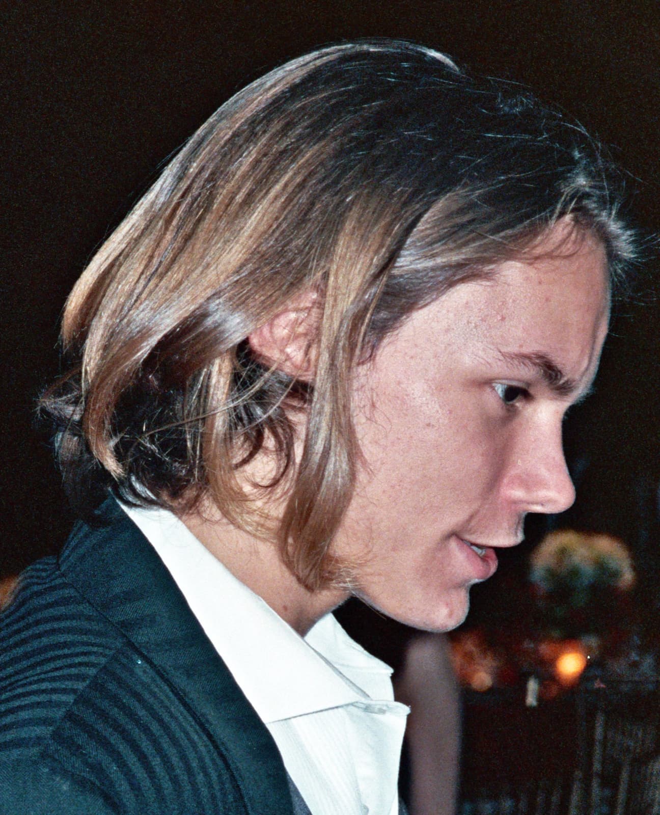 River Phoenix in profile, long hair falling forward, in a dark jacket and white shirt