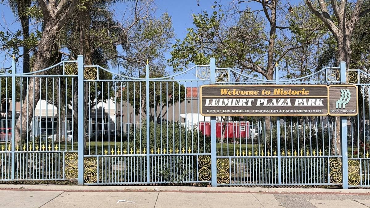 A brown sign reading Welcome to Historic Leimert Plaza Park on an ornamental fence with trees behind