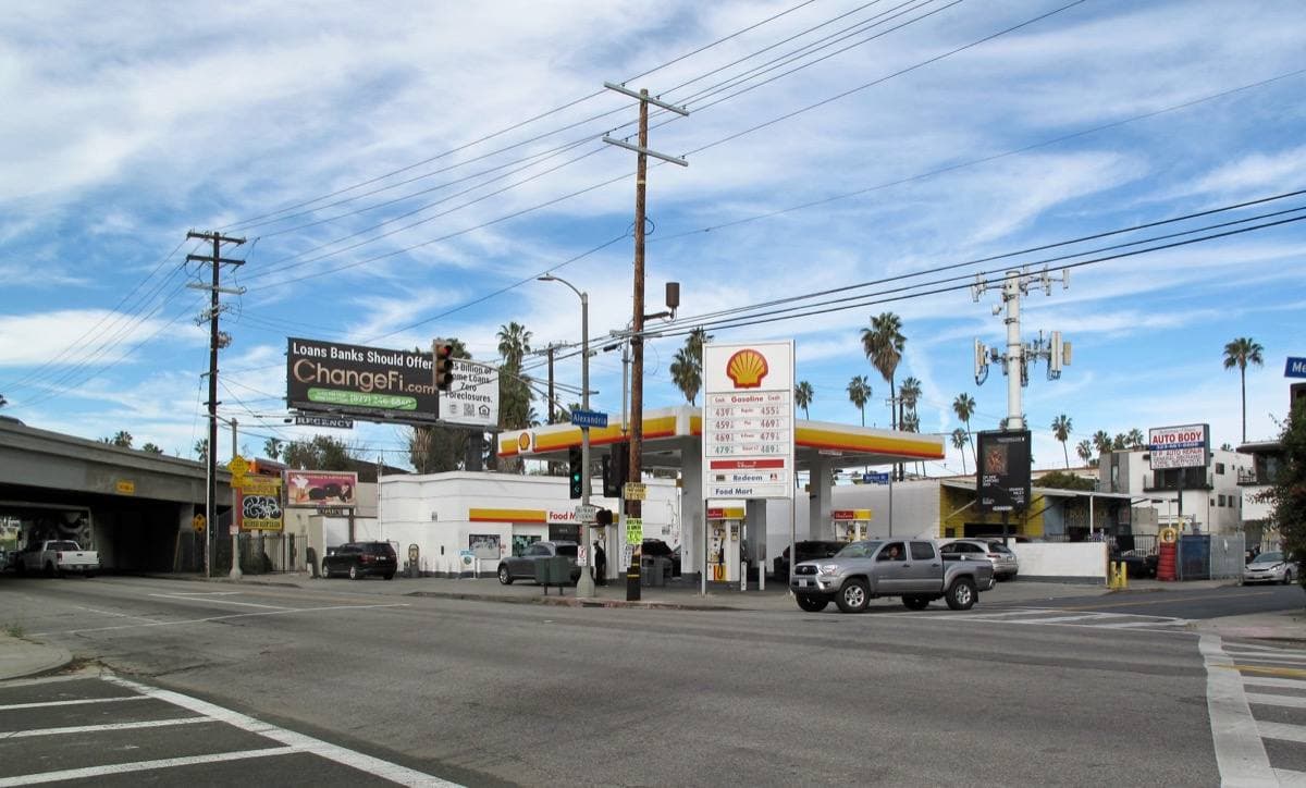 A wide street crossing with a petrol station, power lines, wooden poles and palm trees