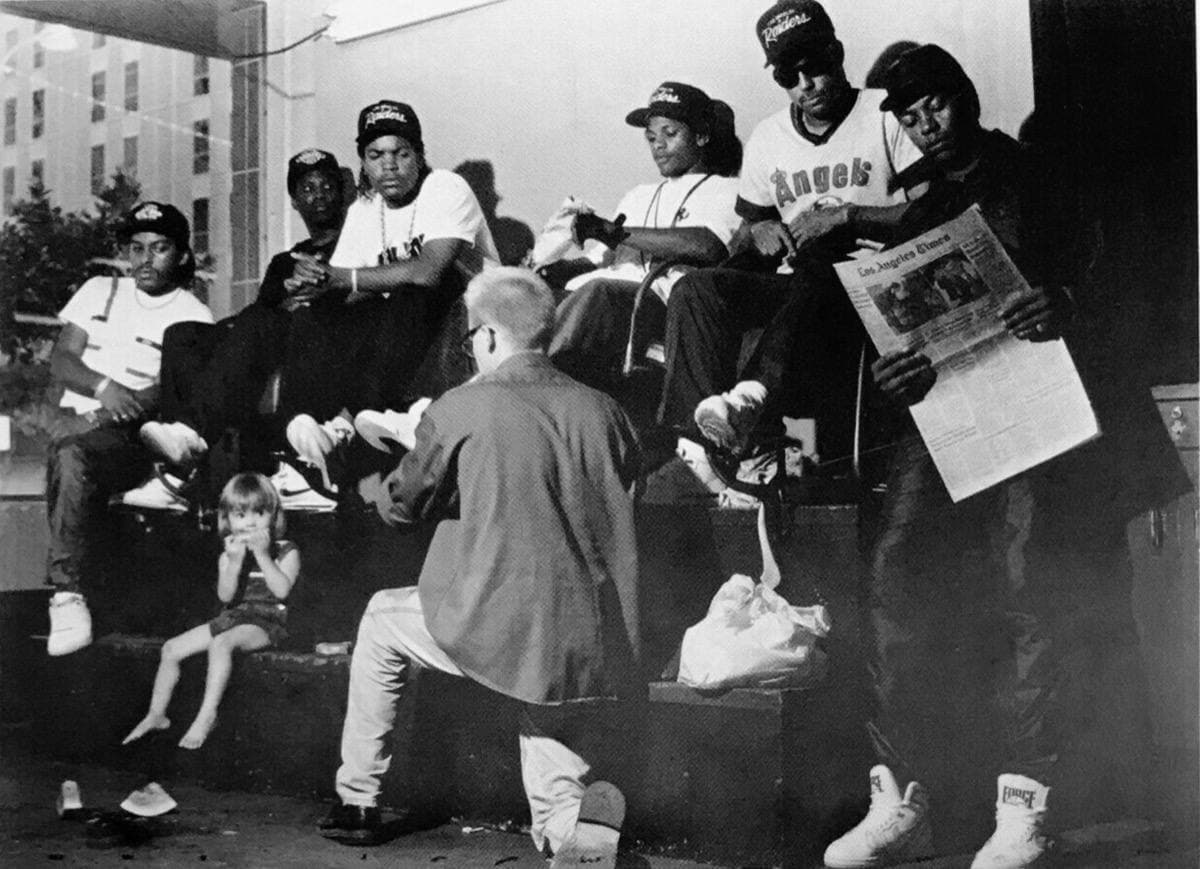 Six young men in caps and dark jackets sitting and crouching together in a black-and-white group photograph