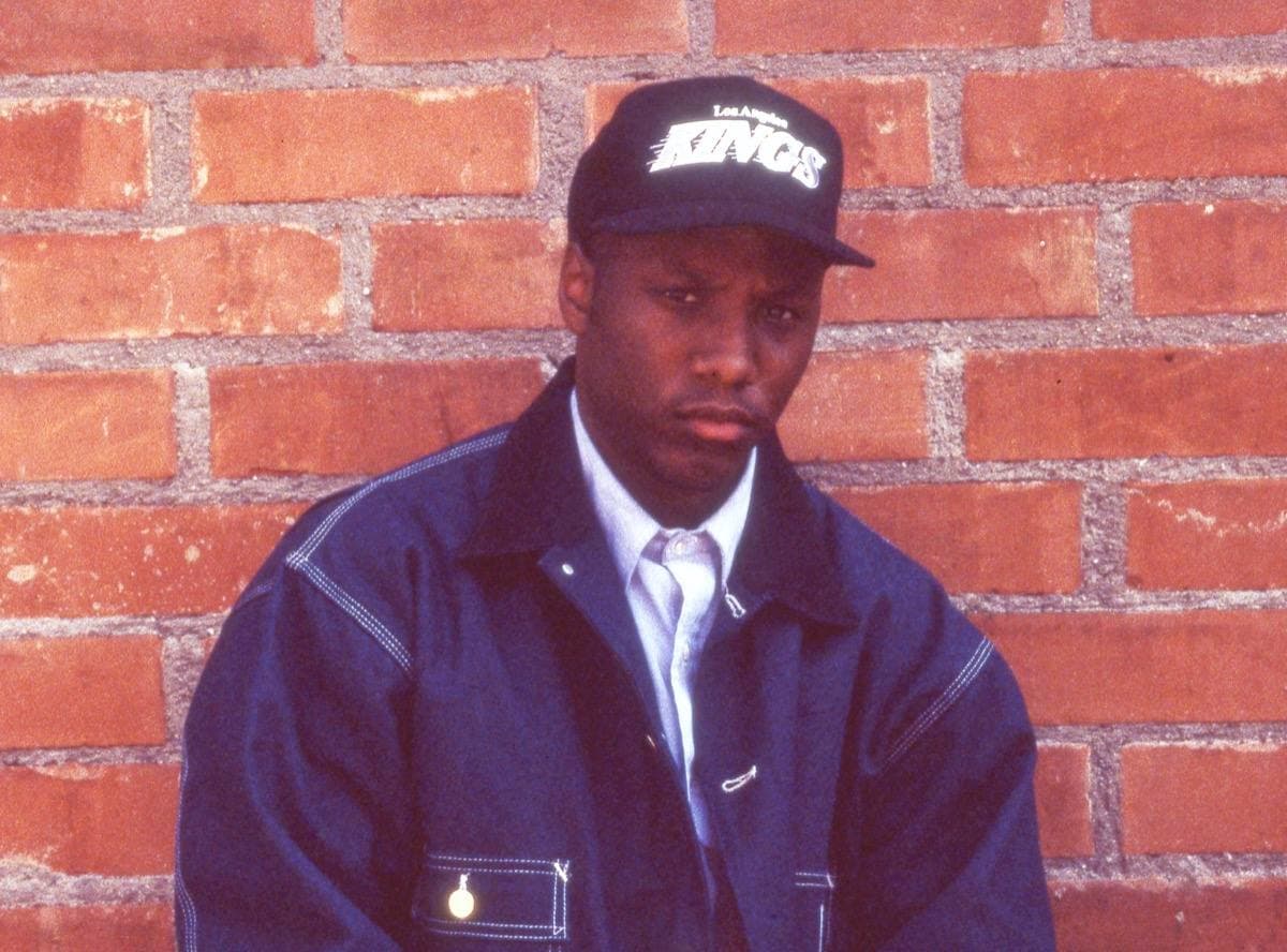 MC Ren in a cap and a dark denim jacket against a red brick wall