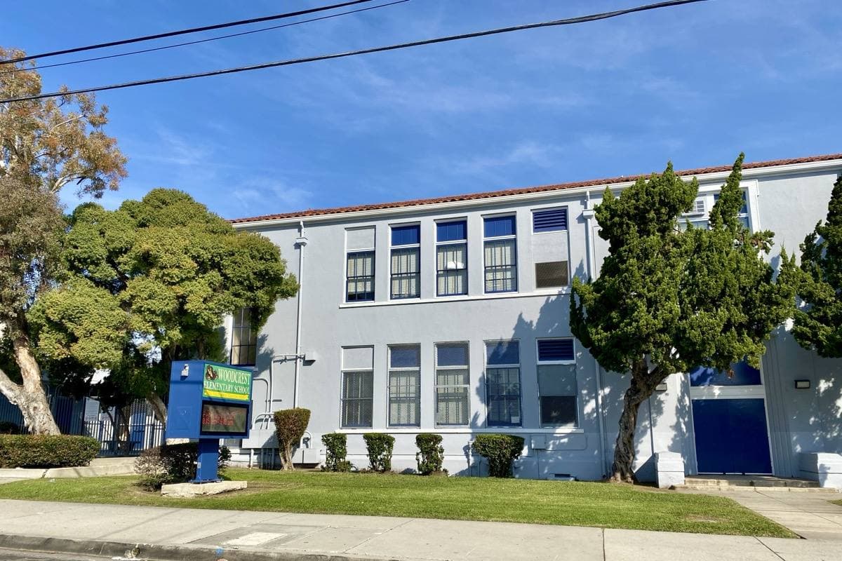 A low school building and a parked car on a quiet street with palm trees and a clear sky