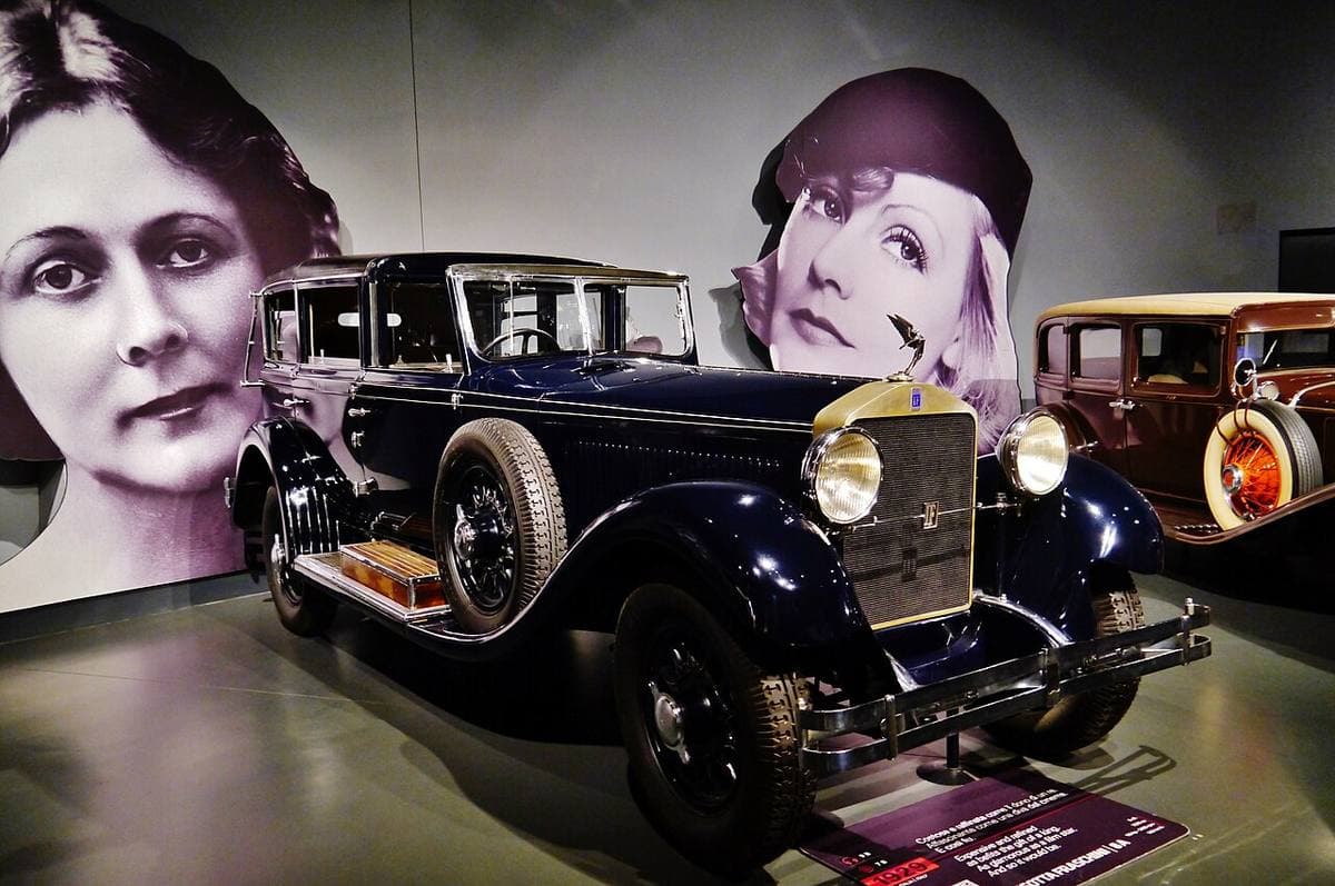A long black 1920s limousine on a museum floor in front of a large image of Gloria Swanson’s face