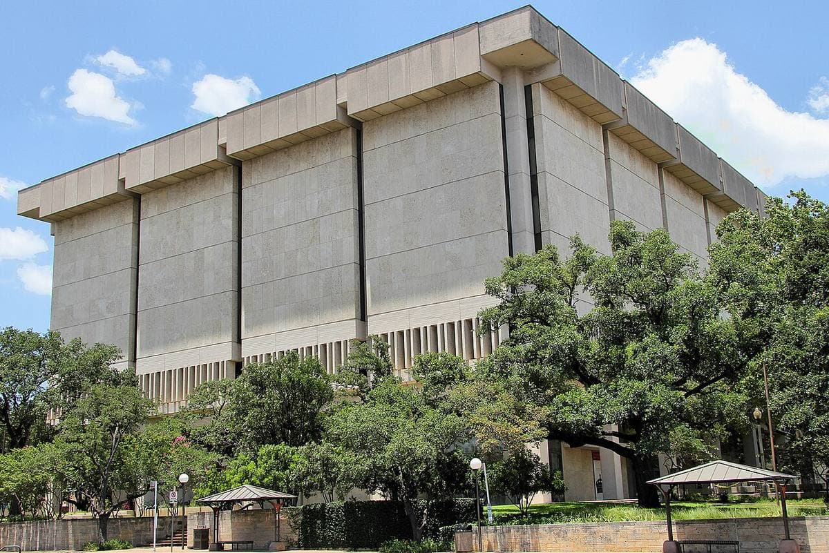 A tall pale stone building with long vertical panels behind oak trees