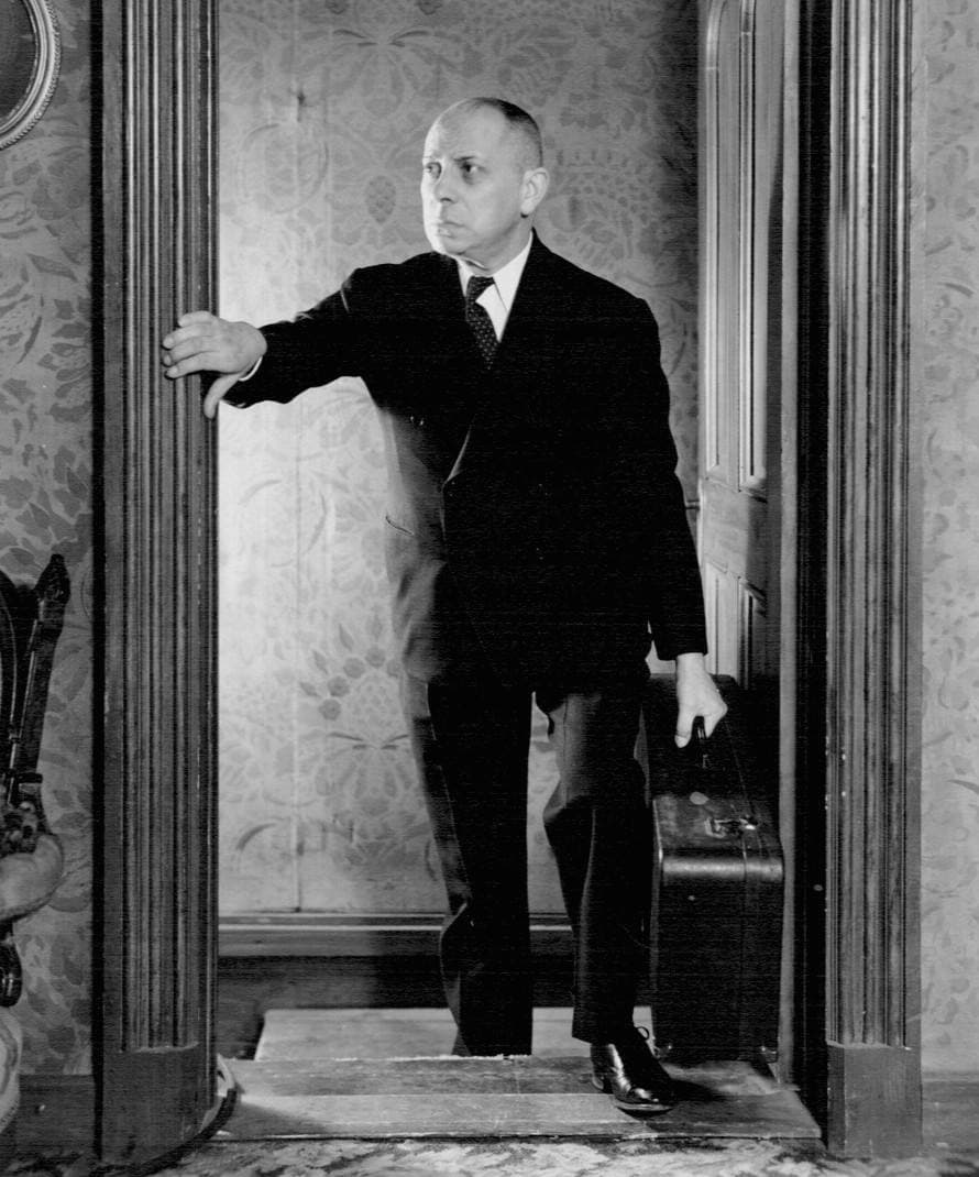 Erich von Stroheim in a dark suit standing in a doorway with a suitcase