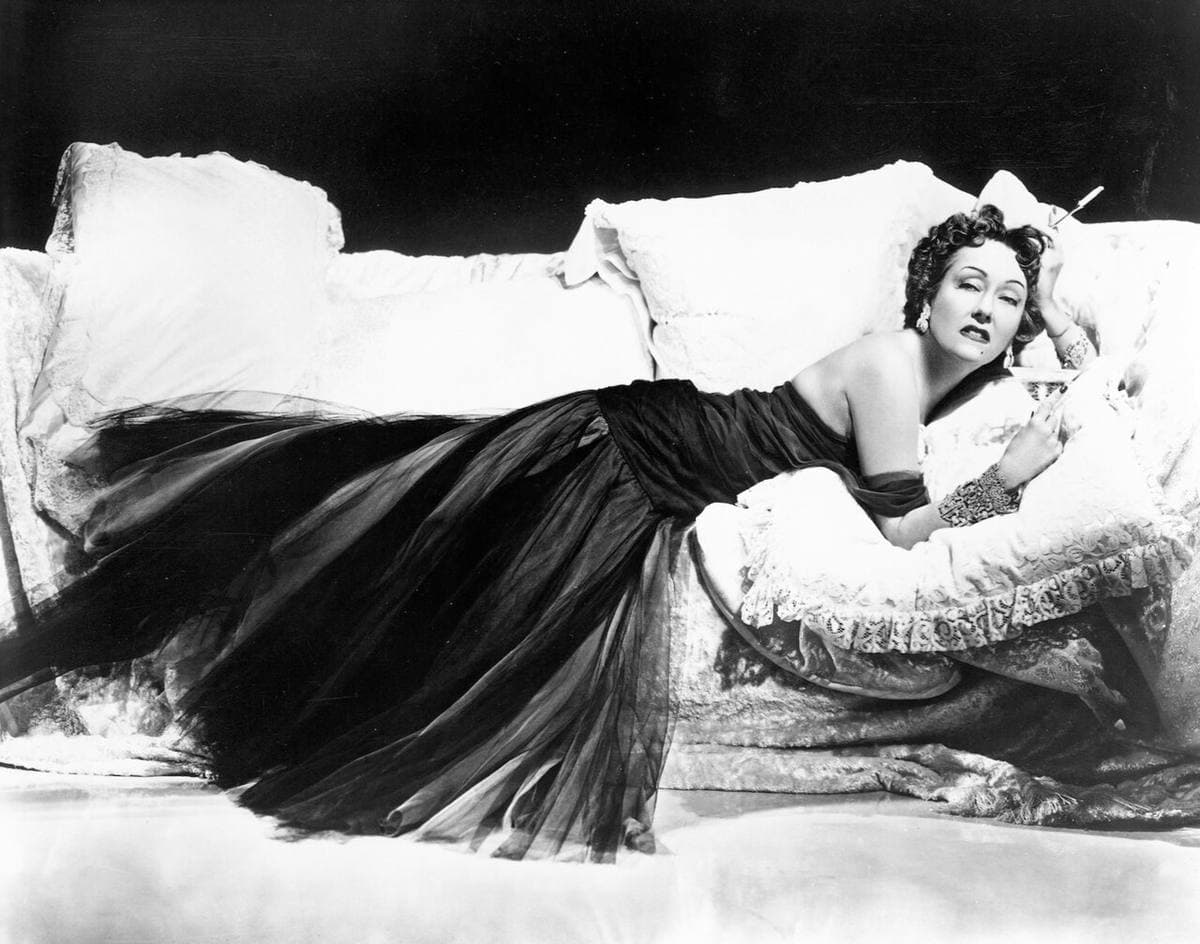 Gloria Swanson in a dark gown reclining on a pale sofa, one arm raised