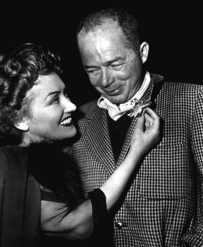 Gloria Swanson, laughing, straightens Billy Wilder’s bow tie; he looks down at her, smiling