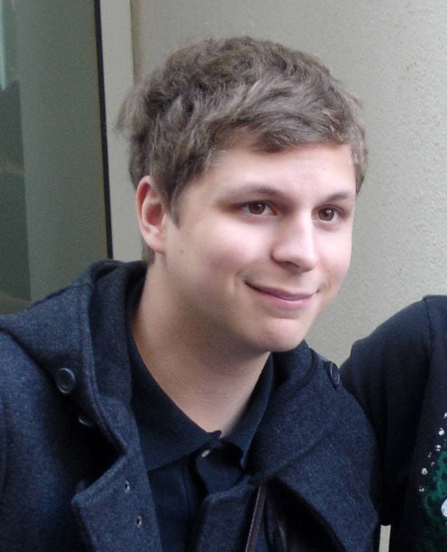 Michael Cera, with a soft fringe, in a navy hooded jacket, smiling sideways