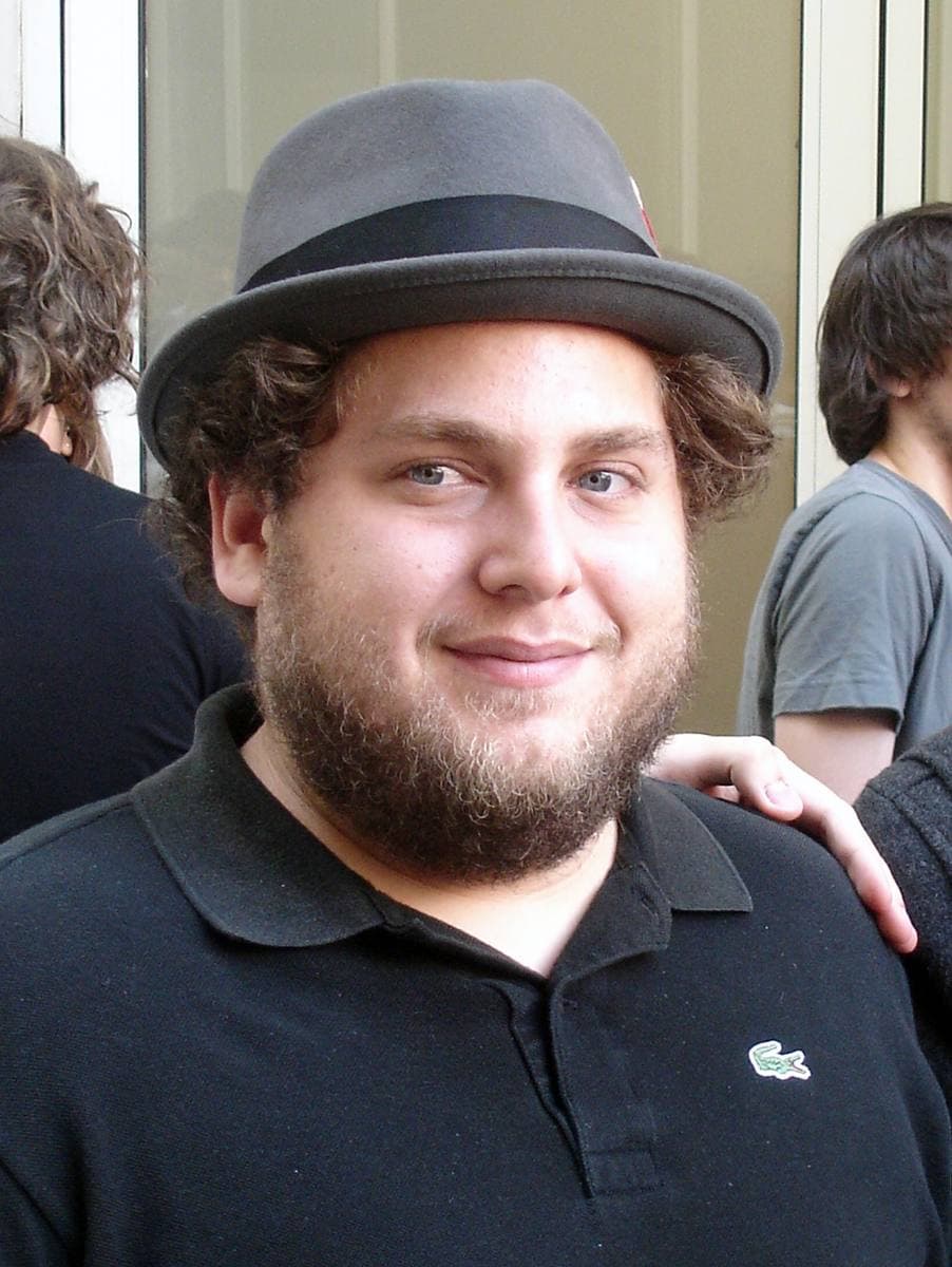 Jonah Hill, bearded, in a grey trilby and black polo shirt, smiling
