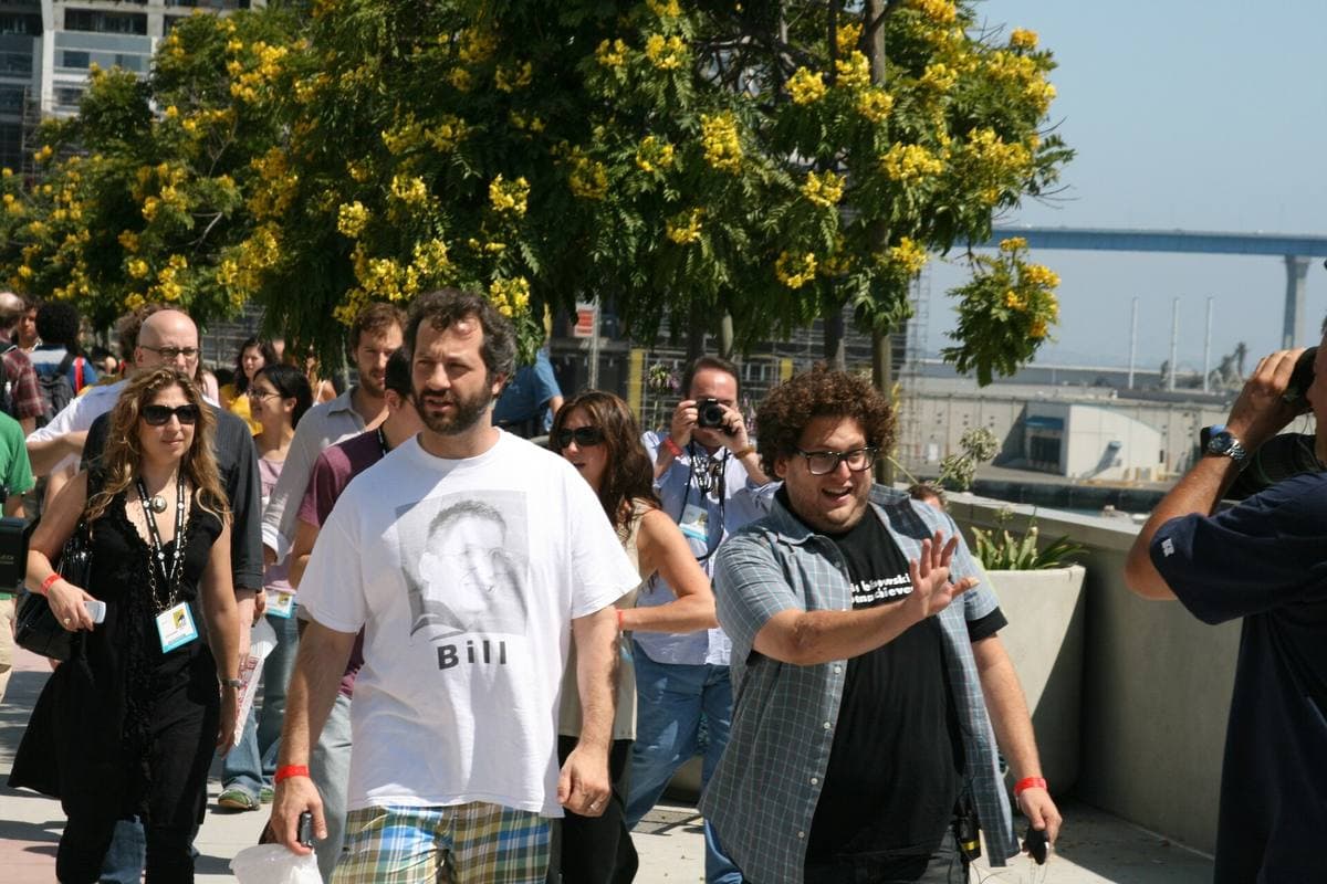 Jonah Hill waving among a crowd outdoors, Judd Apatow in a white T-shirt beside him