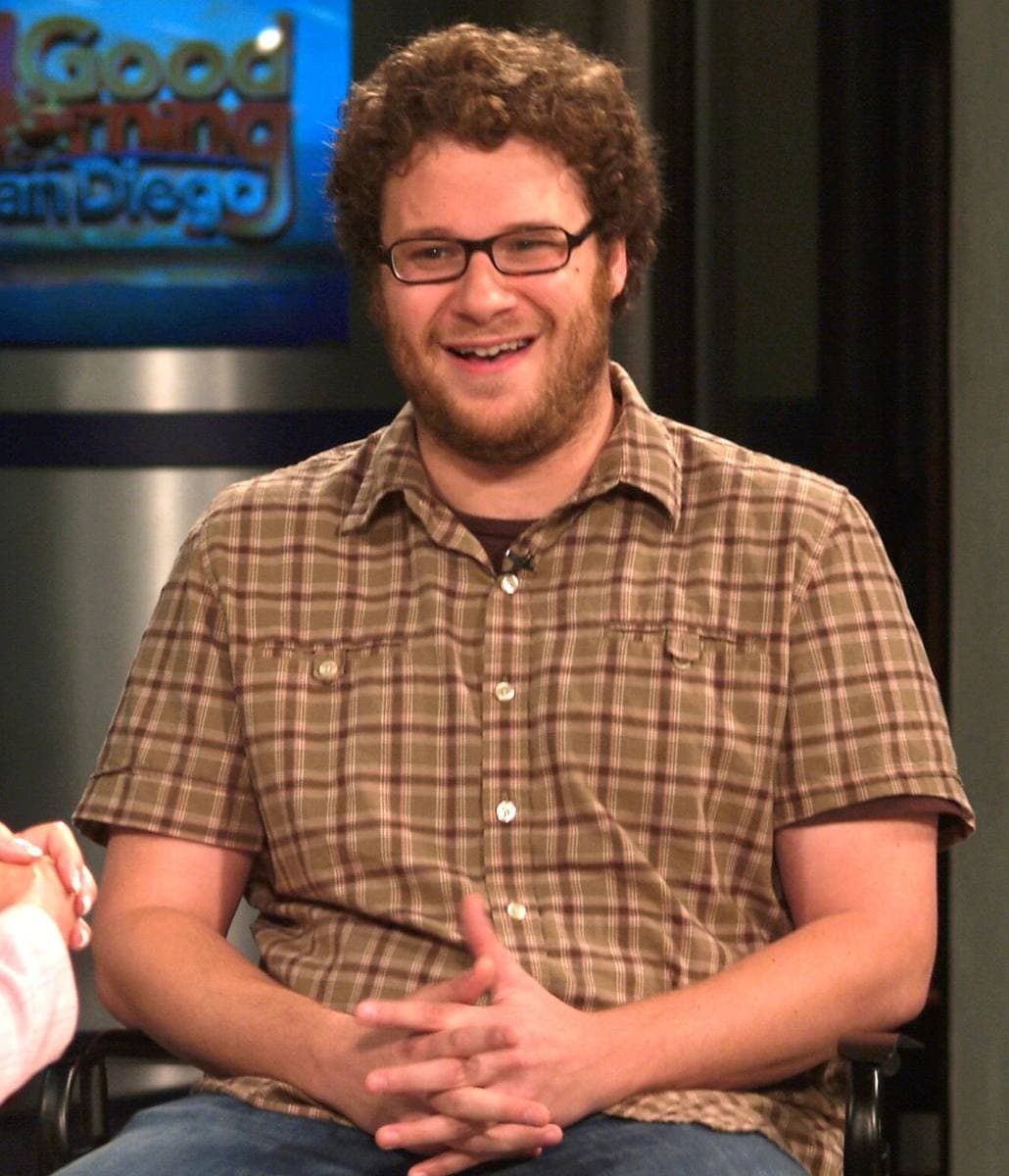 Seth Rogen in a checked shirt and glasses, seated and laughing